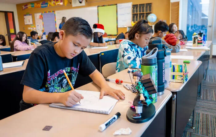 Elementary school students sit at desks in a classroom, focused on writing and drawing in notebooks, with water bottles and school supplies on their desks.